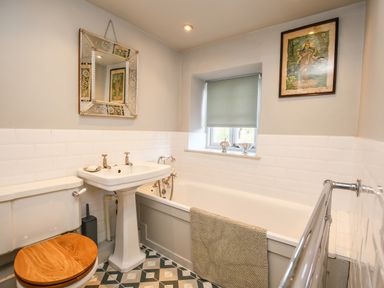 A bathroom with a toilet with wooden seat a pedestal sink a bathtub with a towel a window and framed pictures at The Old Post Office in Coln St Aldwyns