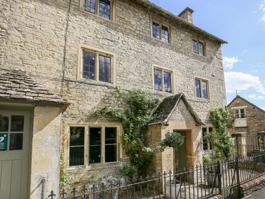 Stone exterior of a multi-story house with windows, a door with a small roof, potted plants, and a metal fence at The Old Post Office in Coln St Aldwyns