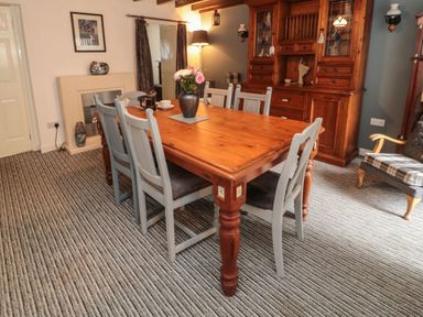 A dining room with a wooden table and six chairs a wooden cabinet and striped carpet at Victoria Cottage in Amble