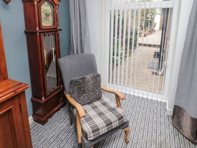 A room with a wooden grandfather clock and an armchair next to a glass door with vertical blinds at Victoria Cottage in Amble