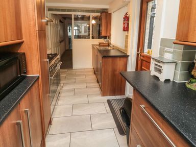 A narrow kitchen with wooden cabinets black countertops a microwave stove sink and a door at Victoria Cottage in Amble