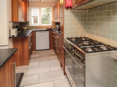 A kitchen with wooden cabinets a gas stove a dishwasher a window with vertical blinds and tiled floor at Victoria Cottage in Amble