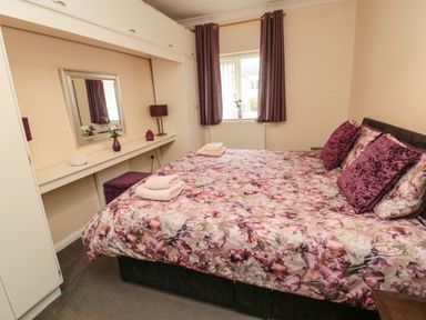 A bedroom with a floral bedspread and purple pillows a desk with a mirror and lamp and a window with purple curtains at Victoria Cottage in Amble