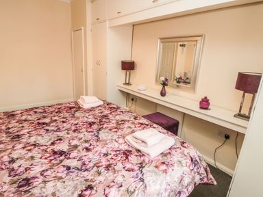 A bedroom with a floral bedspread on the bed towels folded on top a long shelf with a mirror lamp and decorative items at Victoria Cottage in Amble