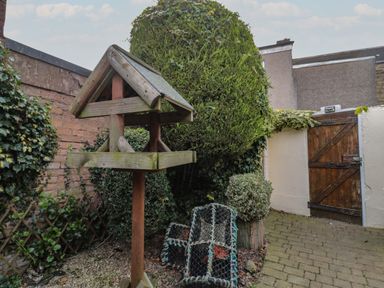 A wooden bird feeder in a garden with a wooden gate and bushes at Victoria Cottage in Amble