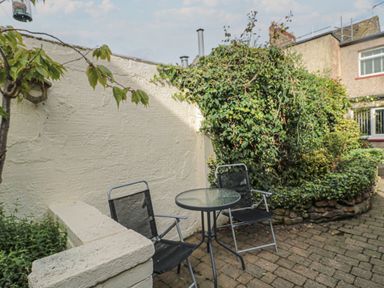 A small outdoor patio with two black chairs and a round glass table near a white wall and green bushes at Victoria Cottage in Amble