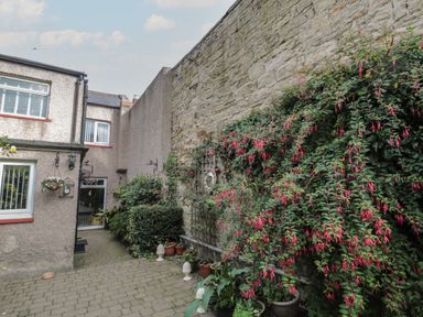 A paved courtyard with potted plants and hanging baskets next to a building and a stone wall with climbing plants at Victoria Cottage in Amble