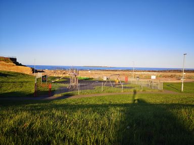 A playground with swings and slides near a grassy area and a sea wall at Victoria Cottage in Amble