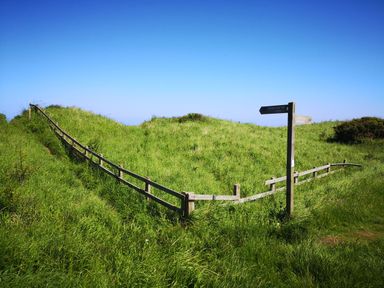A grassy area with a wooden fence and a signpost indicating the England Coast Path at Victoria Cottage in Amble