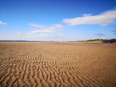 A sandy beach with ripples under a blue sky with some clouds and greenery in the distance at Victoria Cottage in Amble