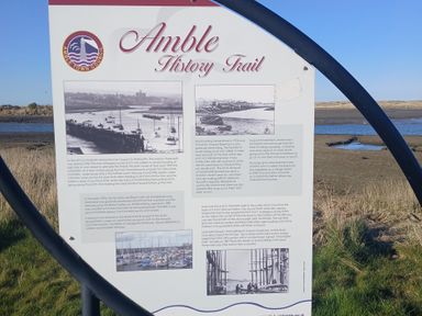 An informational board about Amble History Trail with historical photos and text beside a river at Victoria Cottage in Amble