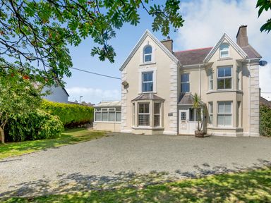 A two story cream-colored house with large windows and a gravel driveway with trees and bushes at Bryn Rhosyn in Nefyn