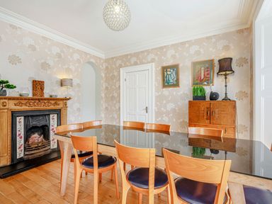 A dining room with a glass table and wooden chairs a wooden fireplace and a cabinet with framed pictures on the wall at Bryn Rhosyn in Nefyn
