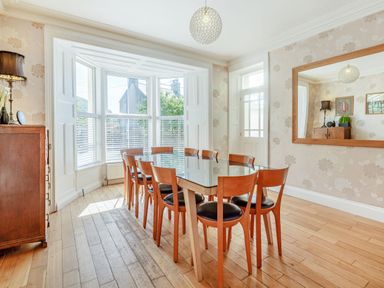 A dining room with a wood table and eight chairs next to a bay window and a wooden cabinet at Bryn Rhosyn in Nefyn