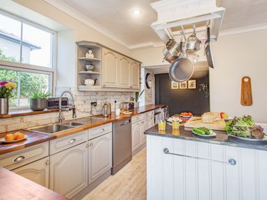 A kitchen with hanging pots and pans a counter with bread vegetables and glasses of juice and cabinets by a window at Bryn Rhosyn in Nefyn