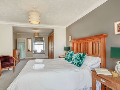 A bedroom with a wooden bed frame white bedding green pillows nightstands with lamps an open book and a window seat with cushions at Bryn Rhosyn in Nefyn
