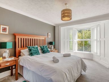 A bedroom with a double bed wooden headboard bedside tables and large bay window with white shutters at Bryn Rhosyn in Nefyn