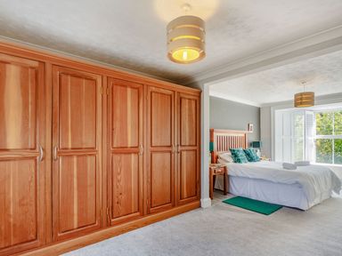 A bedroom with large wooden wardrobes a bed with green pillows and white bed linens and a window with white shutters at Bryn Rhosyn in Nefyn