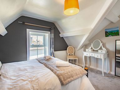 A bedroom with a bed next to a window a chair and a white dressing table with a mirror at Bryn Rhosyn in Nefyn