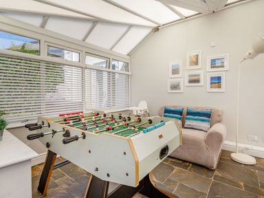 A game room with a foosball table a beige sofa with blue pillows framed pictures on the wall a floor lamp and a high chair at Bryn Rhosyn in Nefyn