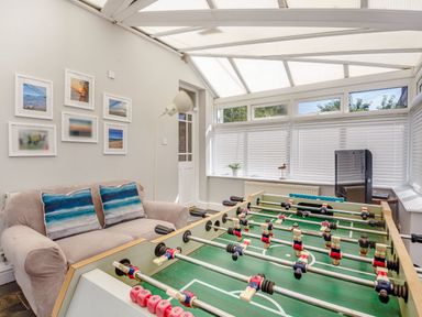 A room with a foosball table a beige sofa with two blue striped cushions framed pictures on the wall and windows with blinds at Bryn Rhosyn in Nefyn