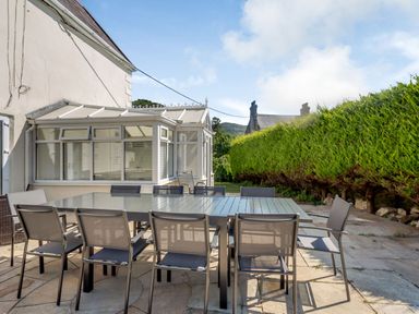 An outdoor patio with a large rectangular table and chairs next to a white building with a conservatory and a hedge at Bryn Rhosyn in Nefyn