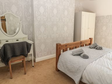 A bedroom with a wooden bed with gray bedding and towels on it a white vanity table with a mirror and a wooden chair with a dark gray blanket and a white wardrobe at Baytree in Alnwick