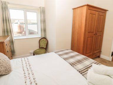 A bedroom with a bed a wooden wardrobe a dresser an upholstered chair and a window with plaid curtains at Baytree in Alnwick