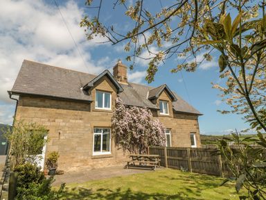 A stone house with a slate roof and flowered vines by a wooden fence and picnic table in a garden at Baytree in Alnwick