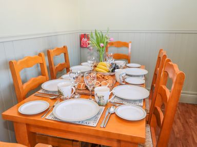 A wooden dining table set with white plates, mugs, glasses, cutlery, bread rolls, bananas, grapes, and a vase of pink tulips at Appletree in Alnwick