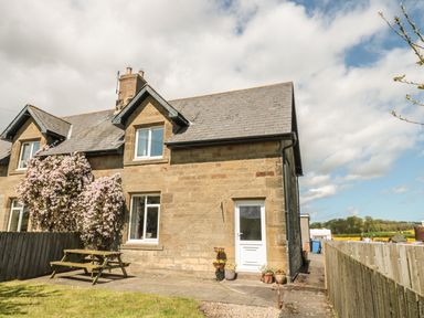 A stone house with a white door and windows with a wooden picnic table and flowering bushes outside at Appletree in Alnwick