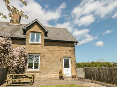 An exterior view of a stone house with two windows a white door a wooden bench and flowering shrub at Appletree in Alnwick