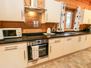 A kitchen with white cabinets black countertops a window with floral curtains a microwave an oven a kettle and a sink at Chloes Lodge in Middleton Ryedale