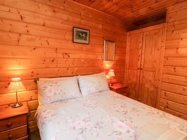A bedroom with wooden walls a double bed with floral bedding two bedside tables with lamps and a wooden wardrobe at Chloes Lodge in Middleton Ryedale
