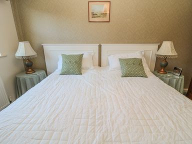 A bedroom with two single beds pushed together white bedcovers green patterned cushions bedside tables with lamps and an alarm clock on a wall with framed artwork at Holly Cottage in Alnwick