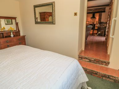 A bedroom with a bed and wooden dresser with flowers leading to a kitchen area at Holly Cottage in Alnwick