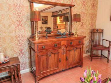 A wooden dresser with mirror and two lamps with plaid shades in a room with patterned wallpaper and a wooden chair at Holly Cottage in Alnwick