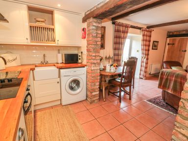 A kitchen area with wooden countertops, a washing machine, a dining table with two chairs, and a living space with a sofa and curtains at Holly Cottage in Alnwick