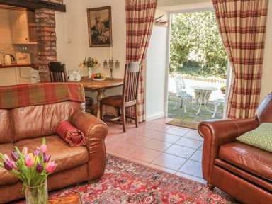 A living room with brown leather sofas a round wooden table with chairs and an open door to a patio at Holly Cottage in Alnwick