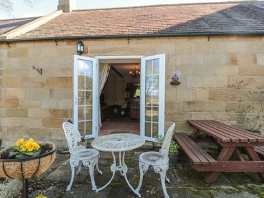 An outdoor patio area with a small round table and two chairs a wooden picnic bench and an open door to a stone building at Holly Cottage in Alnwick