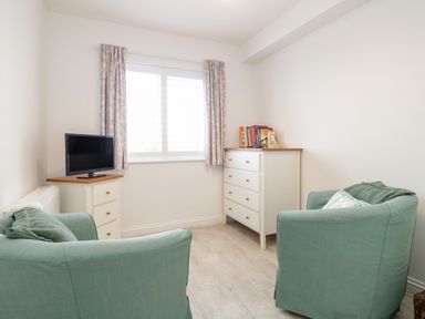 A small room with two green armchairs a white chest of drawers a TV on a corner unit and a window with curtains at 14 Lyme Mews in Seaton Devon