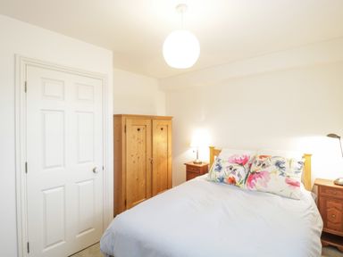 A bedroom with a bed with floral pillowcases two wooden nightstands lamps wooden wardrobe and a white door at 14 Lyme Mews in Seaton Devon
