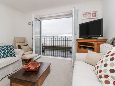 A living room with sofas, armchair, coffee table, television on wooden stand, and open doors with view of the sea at 14 Lyme Mews in Seaton Devon