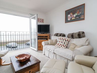 A living room with a sofa, armchair, wooden coffee table, TV on a stand, and an open door to a balcony overlooking the sea at 14 Lyme Mews in Seaton Devon