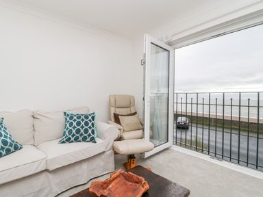 A living room with a white sofa and two blue cushions an armchair and an open door leading to a balcony overlooking the sea at 14 Lyme Mews Seaton Devon