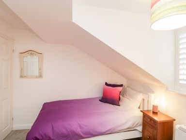 A bedroom with a single bed with purple bedding and two pillows a wooden nightstand with a lamp a mirror on the wall and a window with shutters at 14 Lyme Mews in Seaton Devon