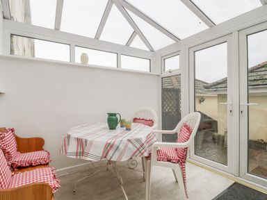A sunroom with a table covered by a plaid tablecloth two chairs with red checkered cushions and glass doors leading outside at 14 Lyme Mews in Seaton Devon