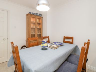 A dining room with a wooden table covered by a blue tablecloth surrounded by wooden chairs and a wooden cabinet with glass doors at 14 Lyme Mews in Seaton Devon