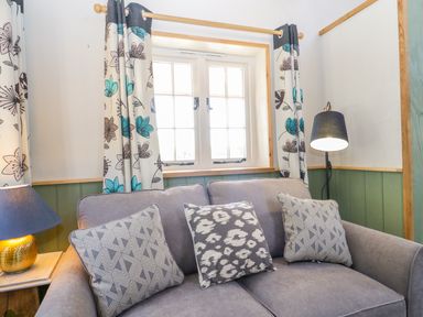 A gray sofa with patterned cushions next to a window with floral curtains and two lamps at Little Willow in Farrington near Child Okeford