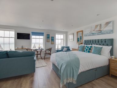 A bedroom with a large bed a teal sofa wooden floor and windows overlooking the sea at 6 The Beaches Weymouth